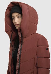 Raudona pūkinė striukė su didele kapišonu, pasižyminti siuvinėtu dizainu, juodu užtrauktuku ir subtiliu logotipo akcentu kairiajame petyje.