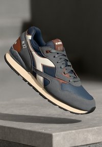 Pantofi sport gri cu un material texturat, având accente albastre și maronii, un logo alb și o talpă de culoare crem. Detalii de design perforate sunt prezente.