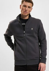 Giacca zip-up grigia scura realizzata in tessuto morbido, con colletto alto, tasche laterali e un piccolo logo sul petto.