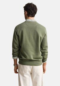 Homme portant un sweat-shirt vert col rond par-dessus une chemise rayée et un pantalon blanc, vu de dos sur fond blanc.