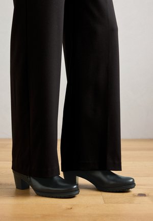 Black heeled shoes with a smooth finish, rounded toe, and textured rubber sole, paired with black wide-leg pants on a wooden floor.