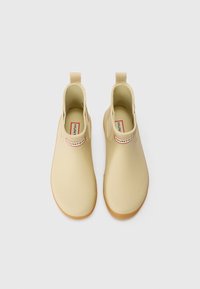 Bottines beiges en caoutchouc, avec une texture lisse, des panneaux latéraux élastiques et une semelle robuste marron. Étiquette du logo en haut.