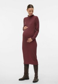 Femme enceinte dans une robe moulante à manches longues de couleur bordeaux, tenant son ventre, portant des bottes noires jusqu'aux genoux, se tenant sur un fond blanc.