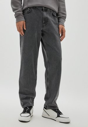 Personne portant un jean gris foncé ample et des baskets blanches avec des accents noirs, debout devant un fond clair uni.