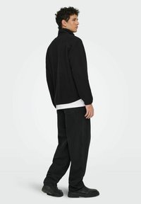 Muškarac stoji bočno, odjeven u crnu flis jaknu, crne hlače, crne cipele i bijelu majicu ispod jakne, uz jednostavnu pozadinu.