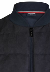 Schwarze Bomberjacke mit Rippkragen, ausgestattet mit einem Reißverschluss vorne und subtiler Steppstruktur. Das Etikett trägt die Aufschrift "HECHTER PARIS."