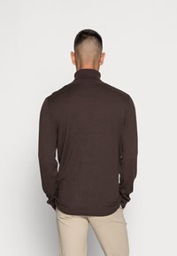 Chemise à manches longues marron foncé avec un col haut. Le tissu semble doux et lisse, présentant un design ajusté et un ourlet légèrement effilé.