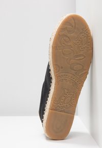 Semelle d'une espadrille noire avec une semelle en caoutchouc texturée affichant un motif floral et le nom de la marque, sur fond blanc.