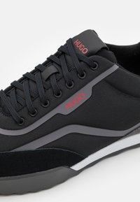 Svart sneaker med ovandel i mesh, en slät mellansula och gummisula. Accentuerad med röd logotyp och grå detaljer. Kraftig design.