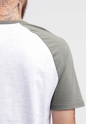 Person iført en hvid skjorte med grå raglanærmer, der viser ryggen og nakken med geometriske tatoveringer.