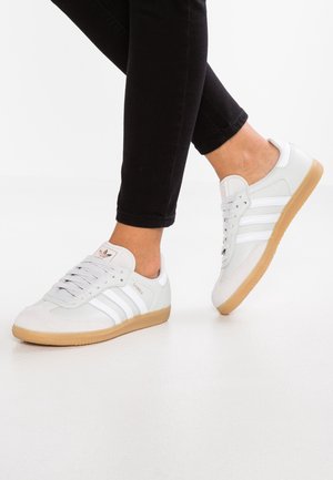 Baskets Adidas Samba blanches à semelles en gomme portées avec un pantalon noir, un pied légèrement levé sur une surface blanche.