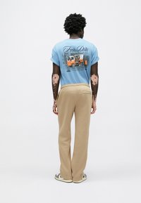 Világoskék pamut póló grafikával a hátulján, bézs puha anyagból készült nadrággal és mintás slip-on sportcipővel párosítva.