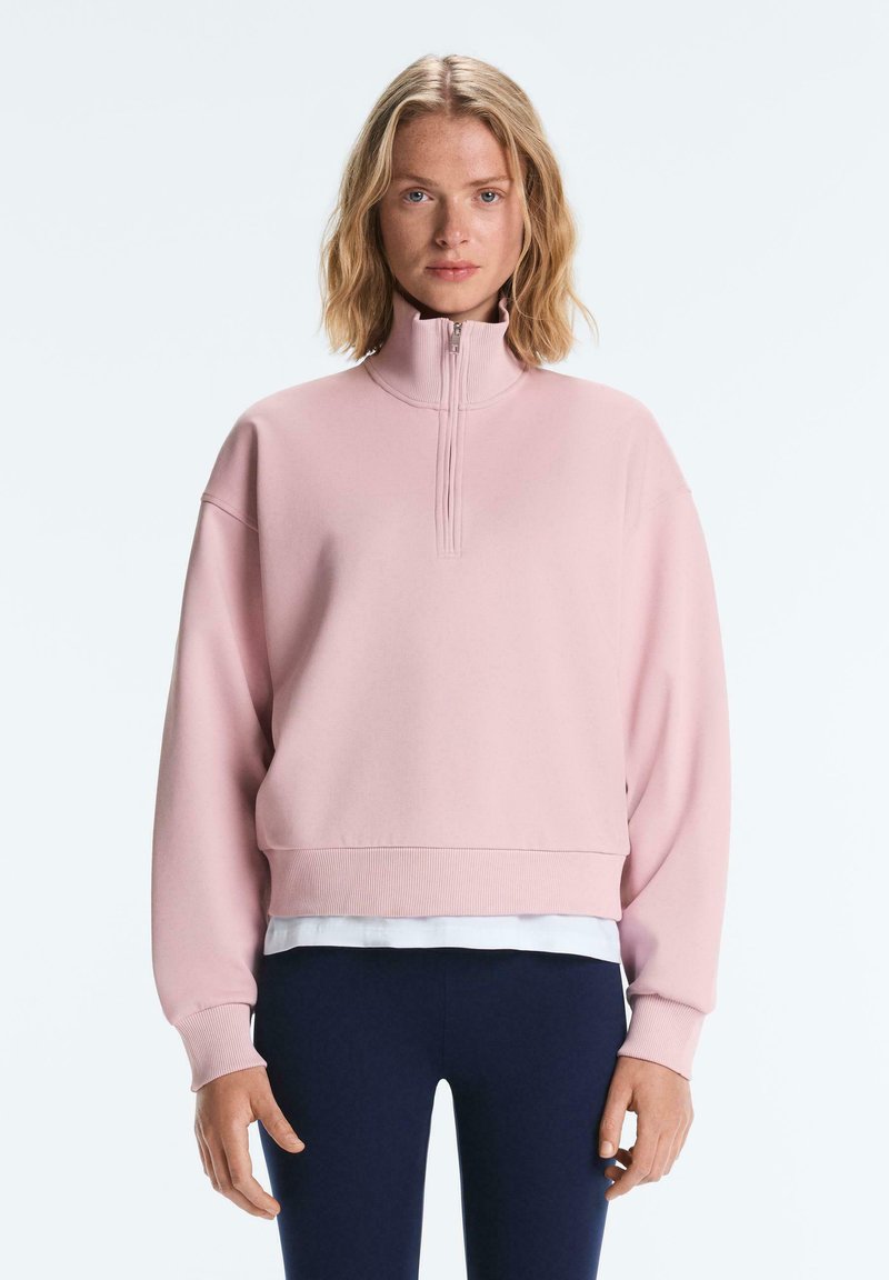 Femme aux cheveux blonds mi-longs portant un sweat-shirt rose clair à fermeture éclair quart de zip, un sous-vêtement blanc et un pantalon bleu foncé, debout sur fond blanc.