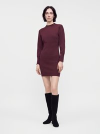Femme debout portant une robe ajustée à manches longues bordeaux foncé et des bottes noires hautes jusqu'aux genoux sur un fond clair uni.