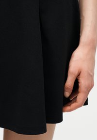 Main droite reposant à côté d'une jupe noire jusqu'aux genoux sur une peau claire, devant un fond uni.