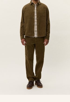 Homme portant une veste et un pantalon en velours côtelé vert olive avec une chemise à carreaux claire en dessous et des chaussures en cuir marron, debout devant un fond uni.