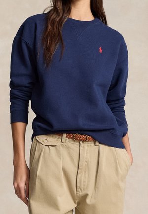 Personne portant un sweat-shirt bleu marine avec un logo rouge et un pantalon plissé beige, les mains dans les poches et une ceinture tressée marron.