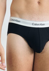 Slip-uri din bumbac negru cu un tiv elastic alb, pe care este înscris logo-ul "Calvin Klein" în negru. Textură netedă și design ajustat.