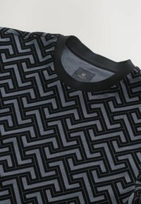 Sort og grå kortærmet sweater med geometrisk mønster med zigzag-linjer, ribbet sort halsudskæring og en subtil struktur. Mærkeetiket synlig.