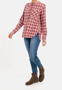 Chemise à carreaux rouge et blanche à boutons, avec un col henley, associée à un jean bleu et des bottines marron. Comprend une poche poitrine et des fentes latérales.