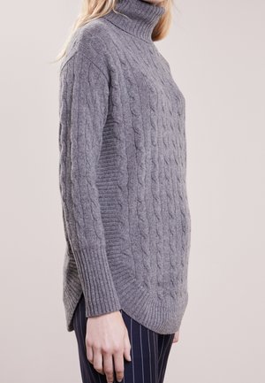 Femme portant un pull gris à col roulé en maille torsadée avec des poignets côtelés et un pantalon bleu marine à fines rayures, vue de profil.