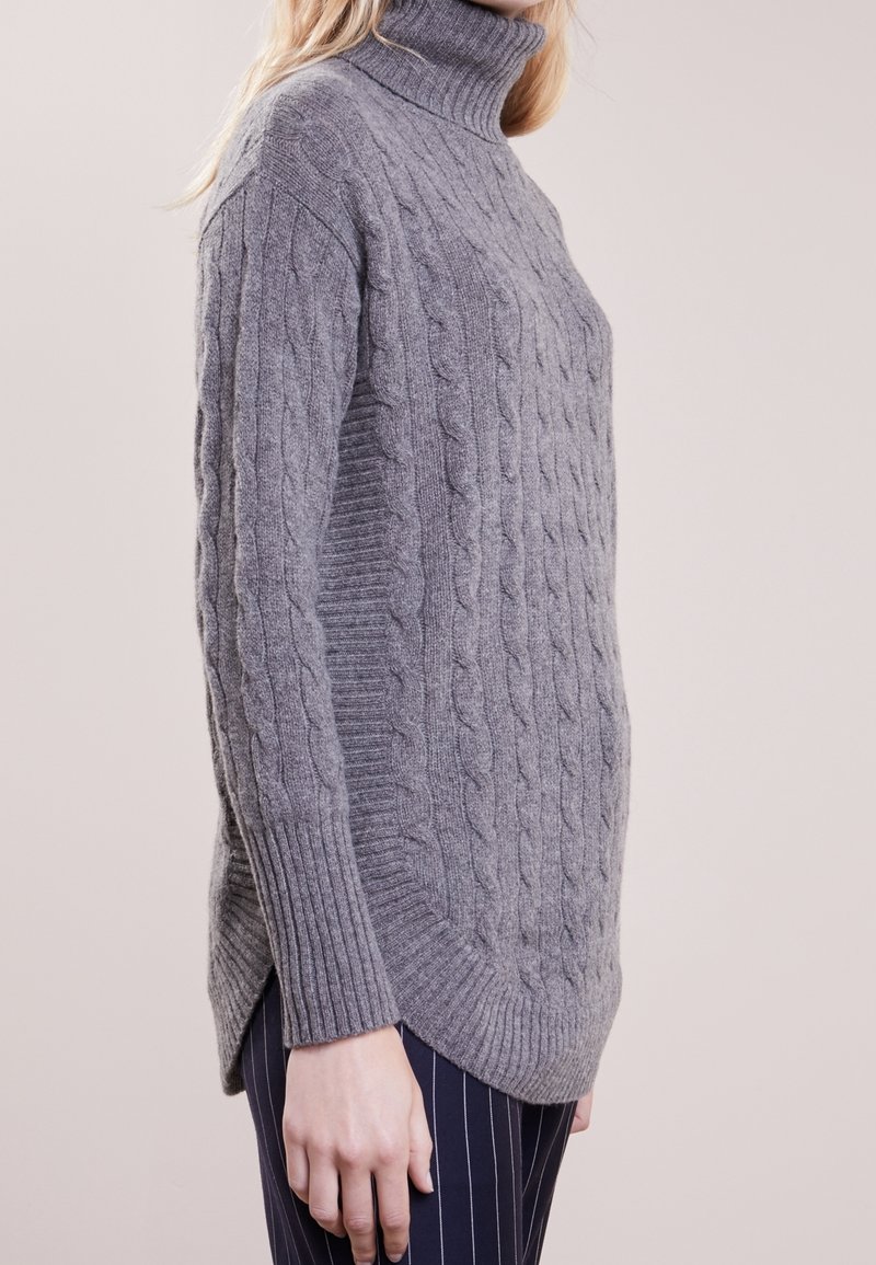 Femme portant un pull gris à col roulé en maille torsadée avec des poignets côtelés et un pantalon bleu marine à fines rayures, vue de profil.