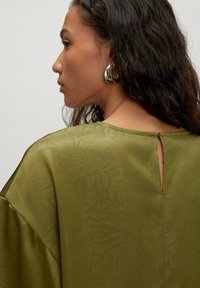 Frau mit welligem, dunklem Haar, die große silberne Creolen trägt und ein olivgrünes, strukturiertes Oberteil mit einem Rückenschlitz und Knopfverschluss anhat.