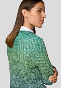 Frau mit glattem braunem Haar, die einen strukturierten grünen Strickpullover über einem weißen Hemd trägt und zur Seite schaut, vor einem hellen Hintergrund.