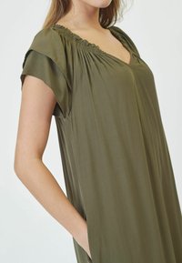 Vestido verde oliva con un corte relajado, que cuenta con mangas cortas y cuello fruncido. Tela suave y lisa con bolsillos laterales.