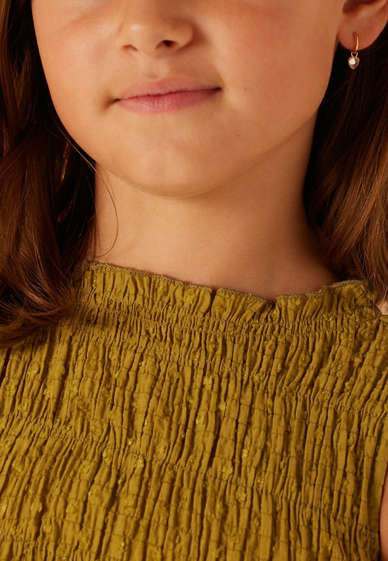 Top smocké texturé jaune avec un col bateau. Présente un tissu froncé et une bordure à volants. Petite boucle d'oreille visible à l'oreille.
