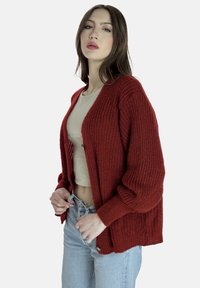 Gebreid vest in dieprood met een ontspannen pasvorm, open voorkant, brede mouwen en geribbelde structuur. Gecombineerd met een licht topje en een high-waisted jeans.