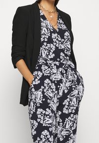 Woman wearing black blazer over navy blue jumpsuit with white floral print, accessorized with layered gold and pearl necklaces.