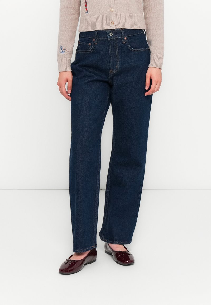 Jeans de pierna recta de color azul oscuro, hechos de denim, con un diseño clásico de cinco bolsillos y costuras visibles. Llevados con bailarinas burdeos.