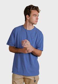 Blaues Baumwoll-T-Shirt mit kurzen Ärmeln, Rundhalsausschnitt und einem dezenten gestickten Design auf der Brust. Getragen mit beigen Hosen.