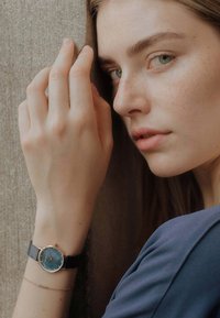Montre ronde avec un cadran bleu et des accents dorés sur un bracelet tissé noir. La main du modèle repose contre un fond texturé.