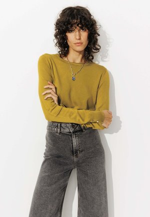 Pull à manches longues couleur moutarde avec une finition texturée et des accents de boutons argentés, associé à un jean noir haut de taille.