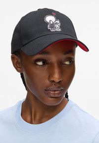 Căciulă neagră cu accente roșii, având un grafic brodat al unei figuri care ține o geantă și o inimă, realizată dintr-un material cu o textură netedă.