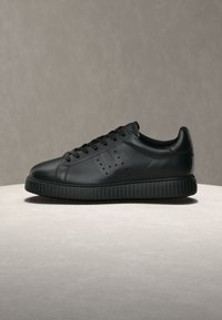 Μαύρο sneaker χαμηλού προφίλ με δέσιμο στο μπροστινό μέρος, διάτρητες λεπτομέρειες στο πλάι και παχύ, υφασμένο πέλμα σε φωτεινή επιφάνεια με μονόχρωμο φόντο.