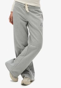 Persona vistiendo pantalones deportivos grises holgados con cintura ajustable con cordón y zapatillas beige, de pie con las piernas cruzadas sobre un fondo blanco.