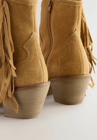 Botas de tobillo de ante en tono marrón con un acabado texturizado, que presentan detalles de flecos y un tacón bloque esculpido. Cierre con cremallera en la parte trasera.
