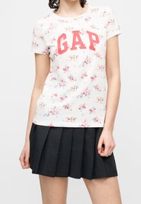 Femme portant un t-shirt blanc fleuri avec des lettres rouges "GAP" et une jupe plissée noire, debout, les mains détendues le long du corps.
