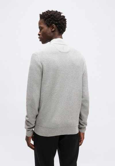 Άνδρας με κοντά σγουρά μαλλιά, φορώντας ανοιχτό γκρι πλεκτό πουλόβερ και μαύρο παντελόνι, γυρισμένος πλάτη σε λευκό ομοιόμορφο φόντο.