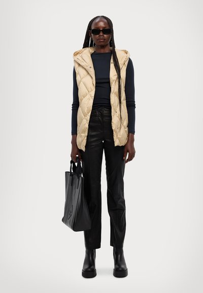 Beige, kviltet ærmeløs vest, sort langærmet top, sorte læderbukser og grove sorte støvler. Holder en sort tote bag med subtilt logo.