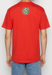 Camiseta de algodón roja con mangas cortas, que presenta un logotipo circular en negro y verde en la parte superior de la espalda. Diseño sencillo, de corte recto.