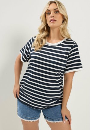 Lyshåret kvinde iført en sort og hvid vandretstribet T-shirt med korte ærmer og blå denims-shorts med blonderkant.