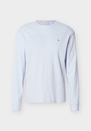 Svetlomodra majica z dolgimi rokavi iz waflaste pletenine z ovratnikom okrog vratu, rebrastimi manšetami in majhno rdečo in belo značko z logotipom "CK" na levi strani prsnega koša.