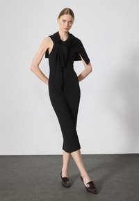 Musta ribbed midi-mekko ilman hihoja, jossa on solmittu musta neule hartioilla ja kiiltävät tummat kengät. Sileä pinta.