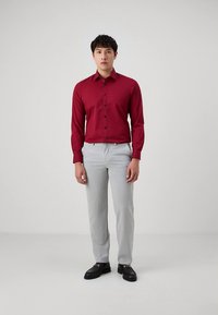 Chemise rouge à manches longues avec une coupe slim, boutons noirs, associée à un pantalon gris clair sur mesure et des chaussures noires. Tissu uni texturé.