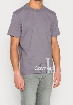 Grå bomulds T-shirt med korte ærmer, rund hals og et stort "Calvin Klein" logo i hvidt nederst, med en subtil struktureret finish.