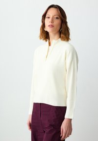 Pull en maille côtelée blanche avec un col montant zippé, doté de manches longues et d'un design court, accompagné d'un pantalon violet foncé.